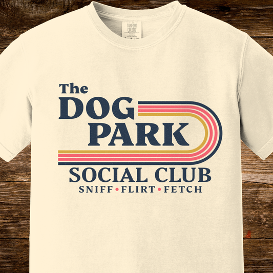 An off-white short-sleeve t-shirt with a retro-style graphic reading 'The DOG PARK SOCIAL CLUB' and 'SNIFF • FLIRT • FETCH' in navy and multicolor stripes, displayed on a rustic dark wood background.