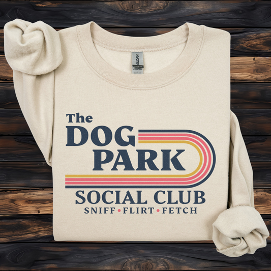 A cream-colored sweatshirt with rolled-up sleeves, featuring a retro-style graphic that reads 'The DOG PARK SOCIAL CLUB' with 'SNIFF • FLIRT • FETCH' underneath. The sweatshirt is displayed flat on a dark wood plank background.