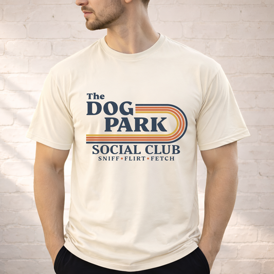 A man wearing a cream-colored t-shirt with a retro-style graphic that reads 'The DOG PARK SOCIAL CLUB' and 'SNIFF - FLIRT - FETCH' in navy blue and orange-yellow stripes, set against a white brick wall background.