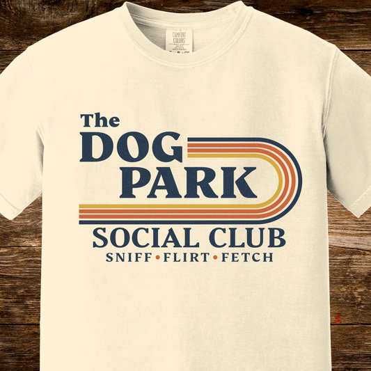 An off-white short-sleeve t-shirt with a retro-style graphic reading 'The DOG PARK SOCIAL CLUB' and 'SNIFF • FLIRT • FETCH' in navy, orange, and yellow stripes, displayed on a rustic dark wood background.