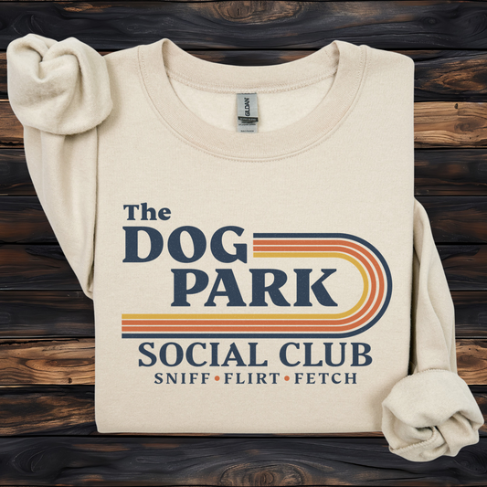 A cream-colored long-sleeve sweatshirt with rolled cuffs, featuring a retro-style graphic that reads 'The DOG PARK SOCIAL CLUB' and 'SNIFF • FLIRT • FETCH' in navy blue and orange-yellow stripes, laid flat on a dark wood background.