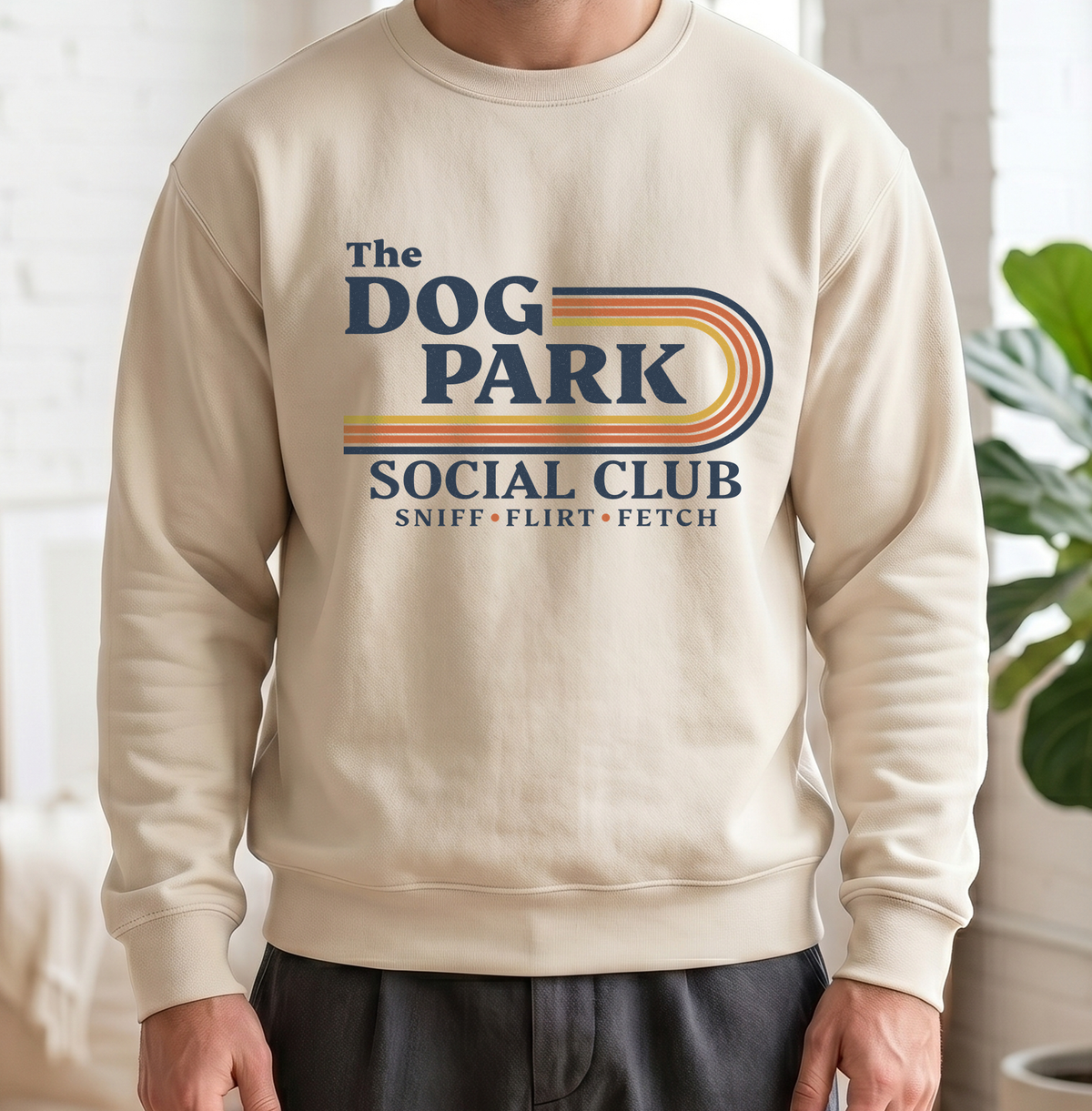 A person wearing a cream-colored crewneck sweatshirt with a retro-style graphic that reads 'The DOG PARK SOCIAL CLUB' and 'SNIFF • FLIRT • FETCH' in navy blue with orange and yellow stripes. The background is a bright, softly blurred indoor setting with a white brick wall and a green plant.