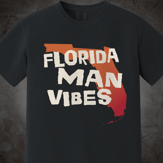 A dark charcoal gray t-shirt featuring a graphic of the state outline of Florida filled with a red-orange gradient and the text 'FLORIDA MAN VIBES' in a distressed white font.