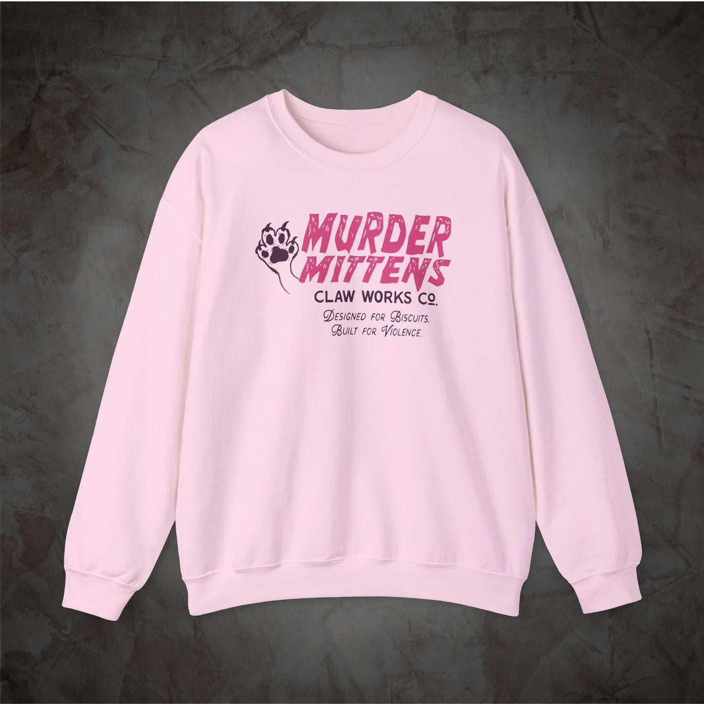 A light pink crewneck sweatshirt featuring a graphic print on the chest with the words 'MURDER MITTENS' in pink and black, a paw print, and 'CLAW WORKS CO.' underneath, displayed against a dark textured background.