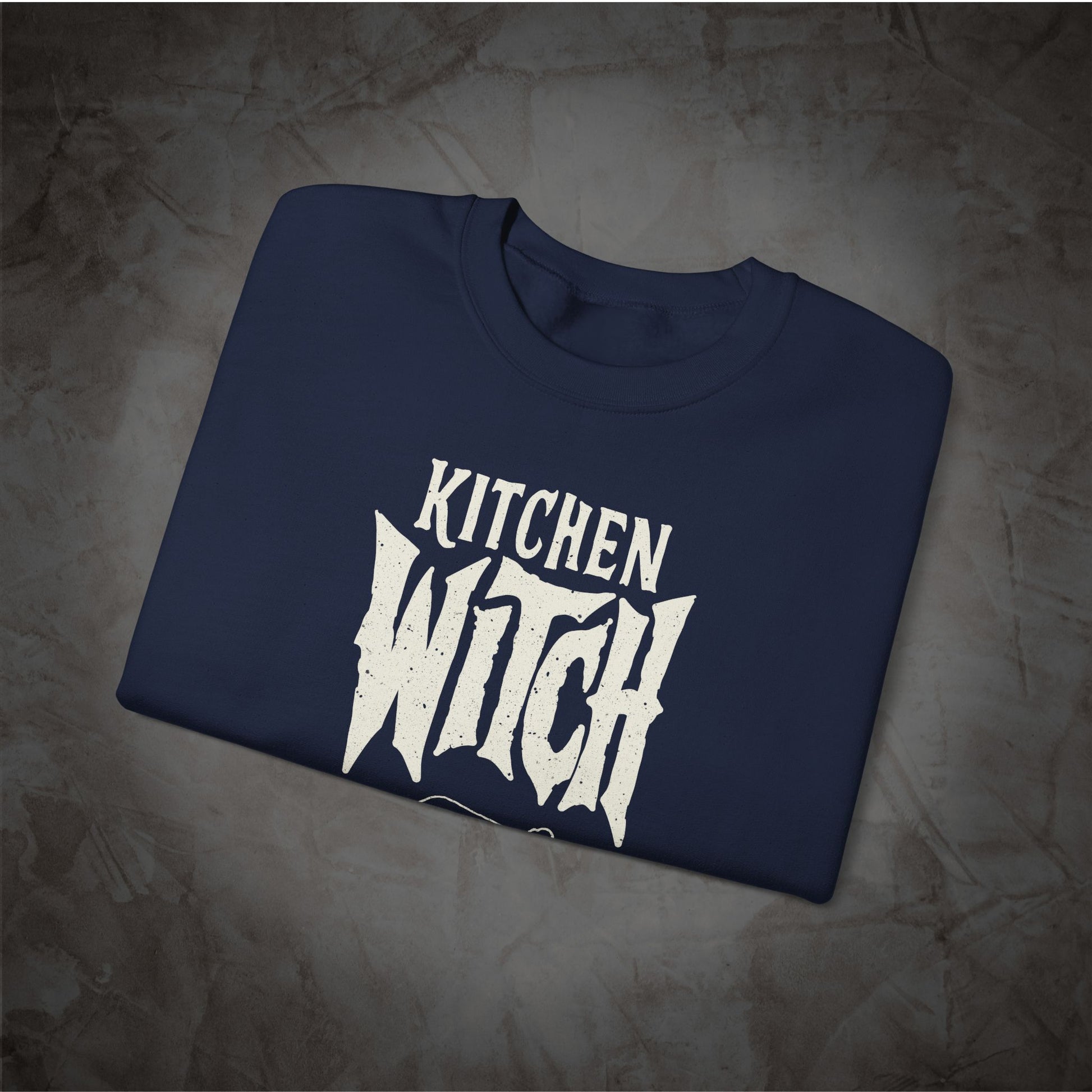 A navy blue folded t-shirt with a distressed white graphic reading 'KITCHEN WITCH' in a stylized font, displayed on a textured gray background.