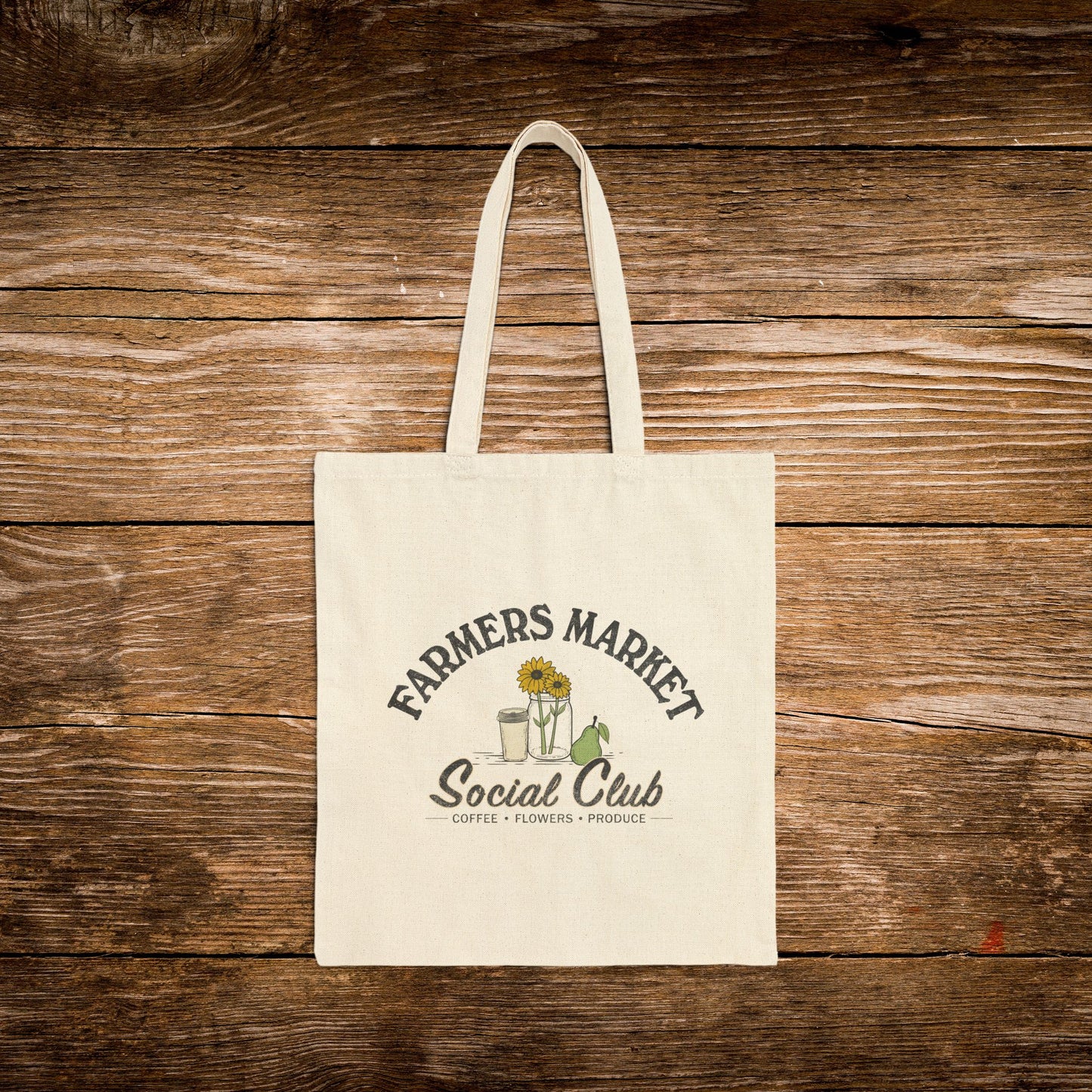 A natural canvas tote bag with a printed graphic reading 'Farmers Market Social Club' and small illustrations of flowers, a sunflower, and a green frog, displayed against a rustic brown wooden plank background.