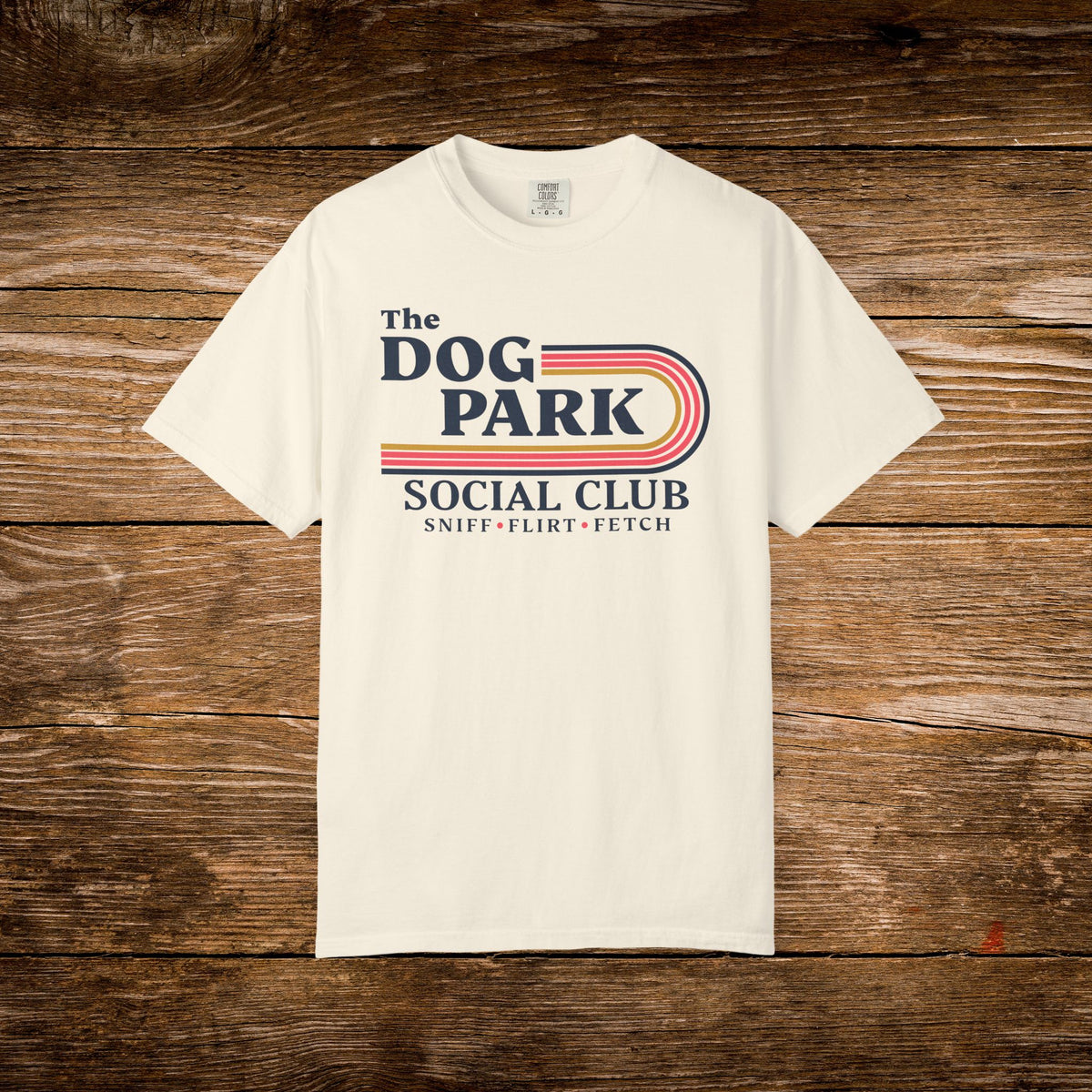 An off-white short-sleeve t-shirt with a retro-style graphic reading 'The DOG PARK SOCIAL CLUB' and 'SNIFF - FLIRT - FETCH' in navy and red, laid flat on a rustic brown wooden background.