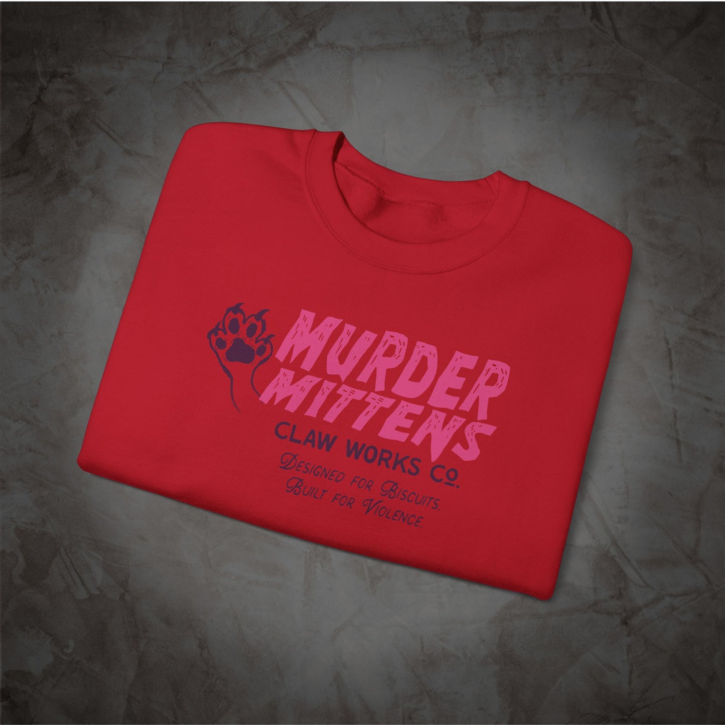 A folded red t-shirt with a pink and dark purple graphic print reading 'MURDER MITTENS' and 'CLAW WORKS Co.' on a textured gray background.
