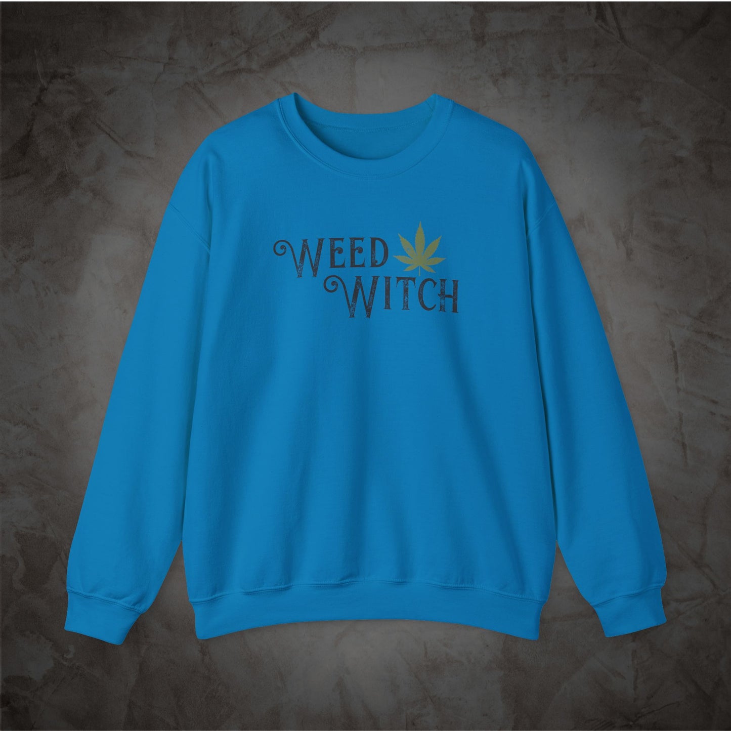 A bright blue crewneck sweatshirt featuring the words 'WEED WITCH' in a dark gray gothic font with a small yellow-green cannabis leaf graphic, displayed on a textured dark gray background.