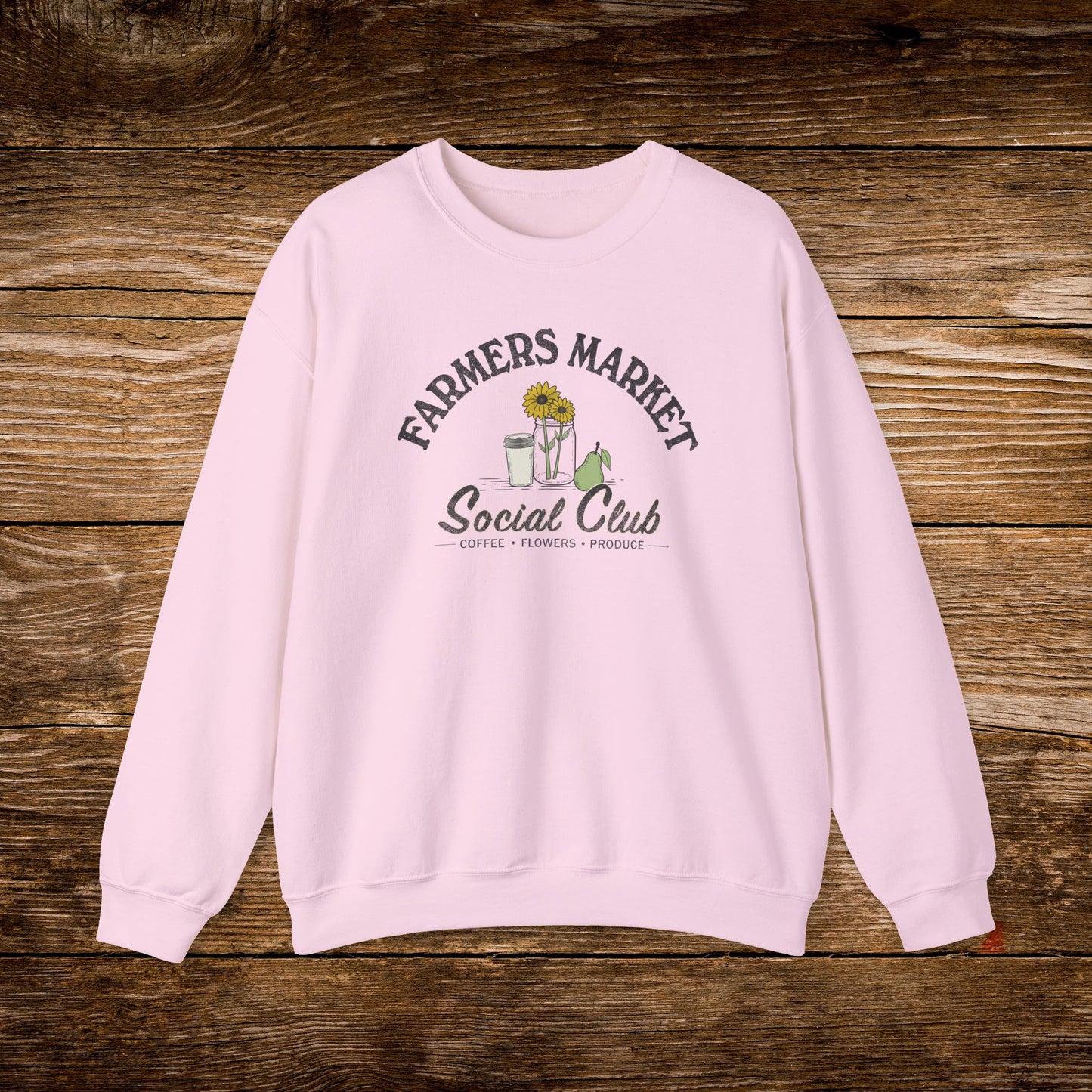 A light pink crewneck sweatshirt with a 'Farmers Market Social Club' graphic on the chest, displayed flat on a rustic wooden plank background.