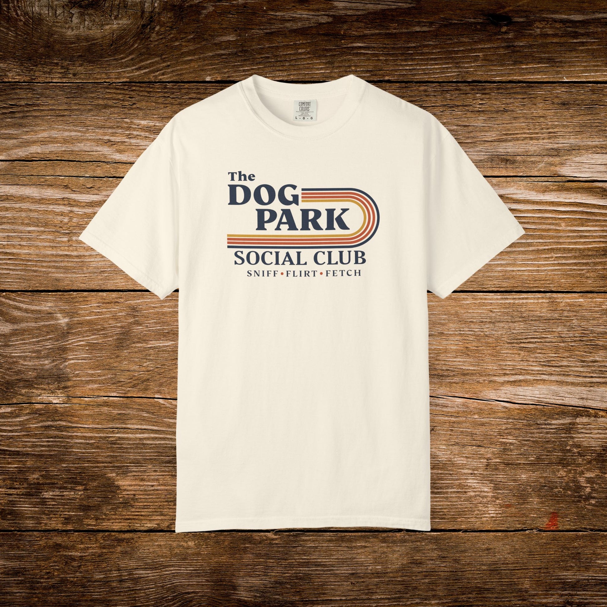 An off-white crewneck t-shirt with a retro-style graphic reading 'The DOG PARK SOCIAL CLUB' and 'SNIFF - FEEL - FETCH' in navy and orange, displayed flat against a rustic brown wooden plank background. The design text reads "Sniff, Flirt, Fetch."