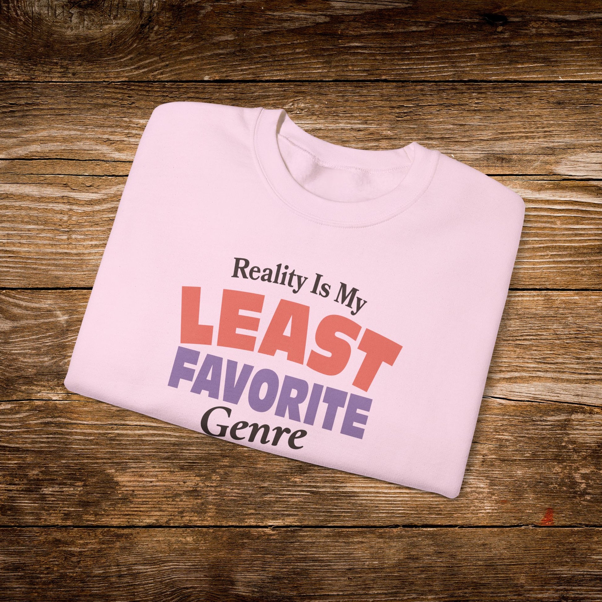 A light pink folded t-shirt with the text 'Reality Is My LEAST FAVORITE Genre' printed on the front, displayed on a rustic brown wooden background.