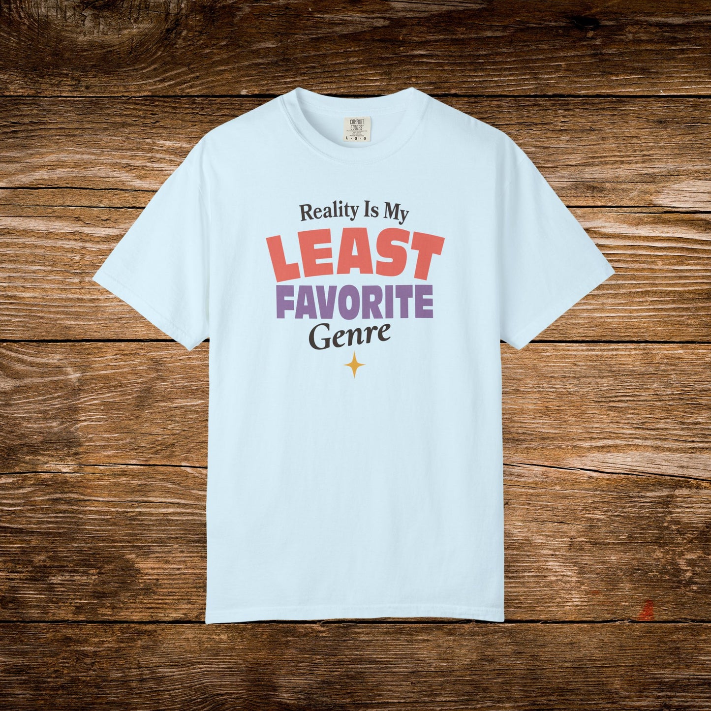 A light blue short-sleeve t-shirt with a graphic text print reading 'Reality Is My LEAST FAVORITE Genre' in red, purple, black, and gold, displayed flat on a rustic brown wooden background.