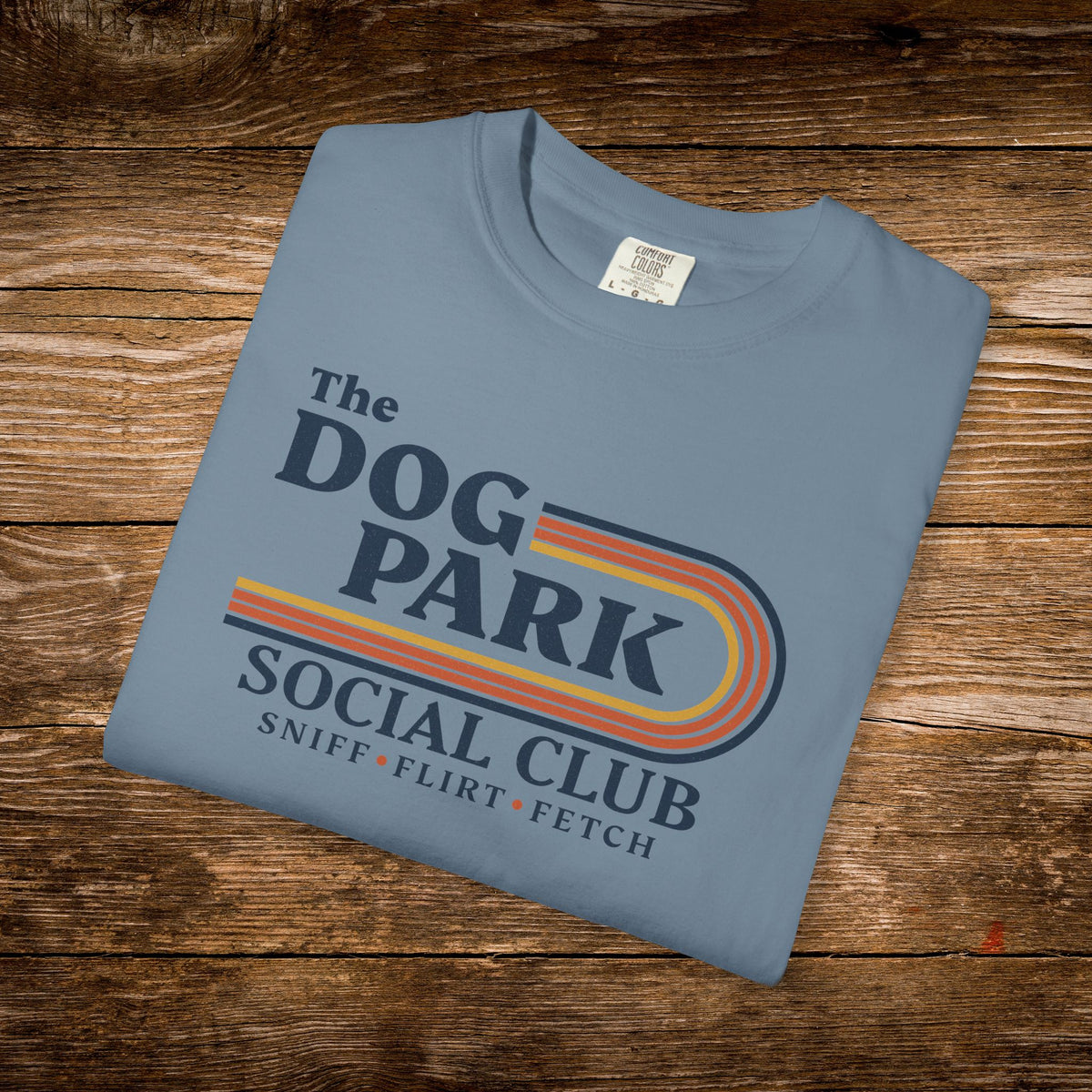 A folded light blue t-shirt with a retro-style graphic reading 'The DOG PARK SOCIAL CLUB' and 'SNIFF • FLIRT • FETCH' in dark blue text with orange and yellow stripe accents, displayed on a rustic wooden background.