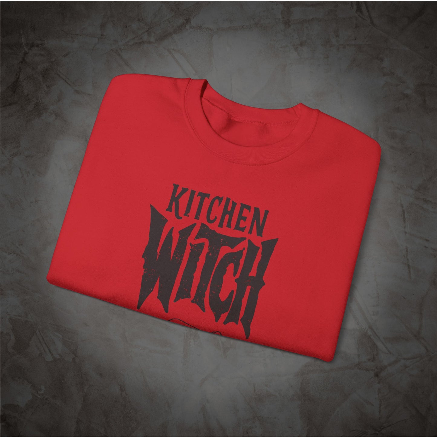 A folded red crewneck t-shirt with a black graphic print reading 'KITCHEN WITCH' in a stylized font, displayed on a textured gray background.