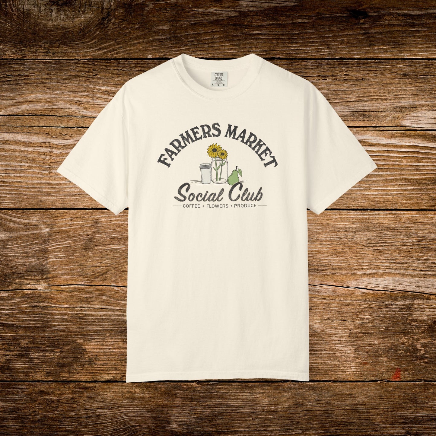 A person lying on grass outdoors holds a blue book over their face, wearing a cream graphic T-shirt that reads 'Farmers Market Social Club' with sunflower and coffee cup artwork, paired with light blue jeans.