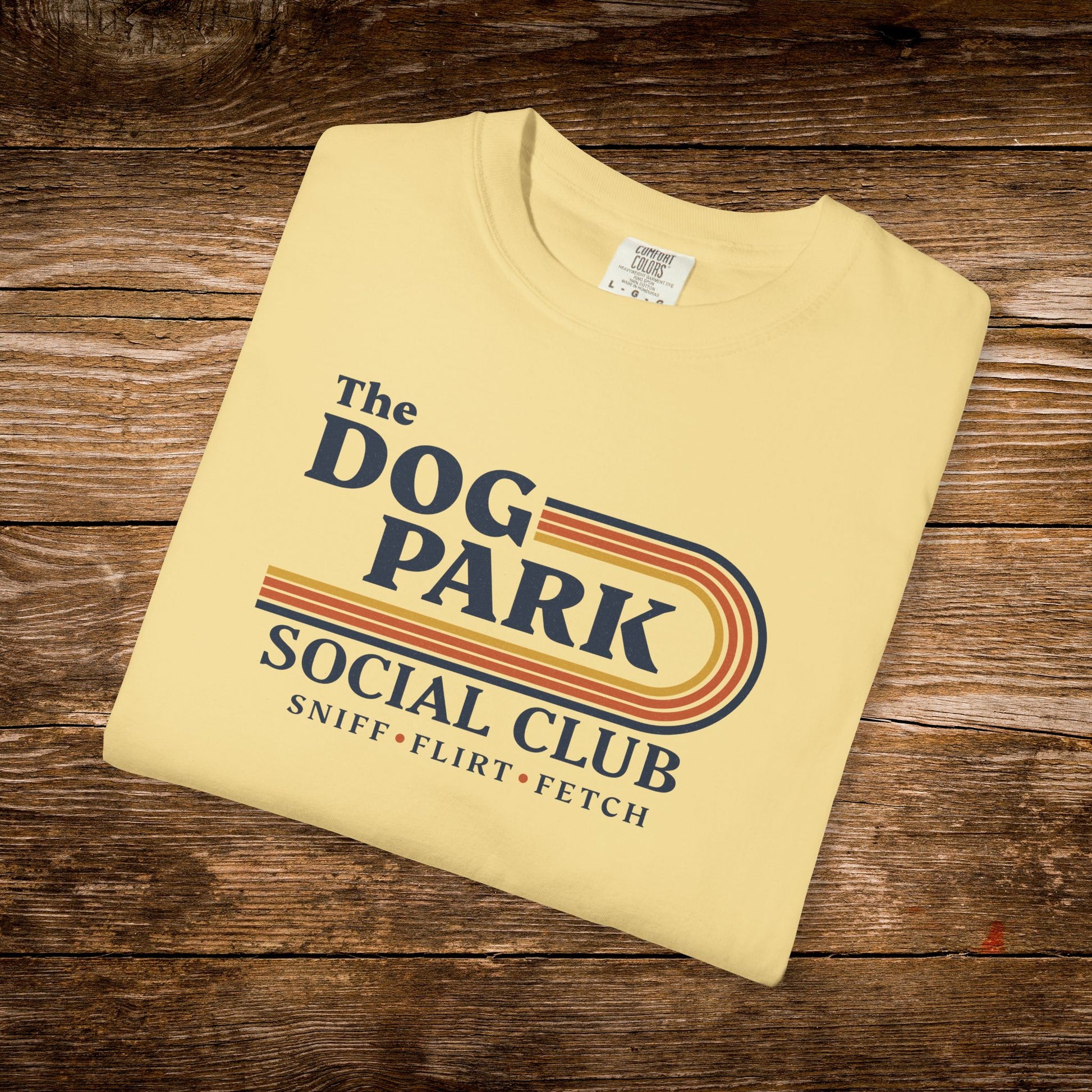 A folded pale yellow graphic T-shirt on a rustic wooden surface. The shirt features a retro-style printed design that reads 'The DOG PARK SOCIAL CLUB' with 'SNIFF • FLIRT • FETCH' underneath. The design text reads "Sniff, Flirt, Fetch."