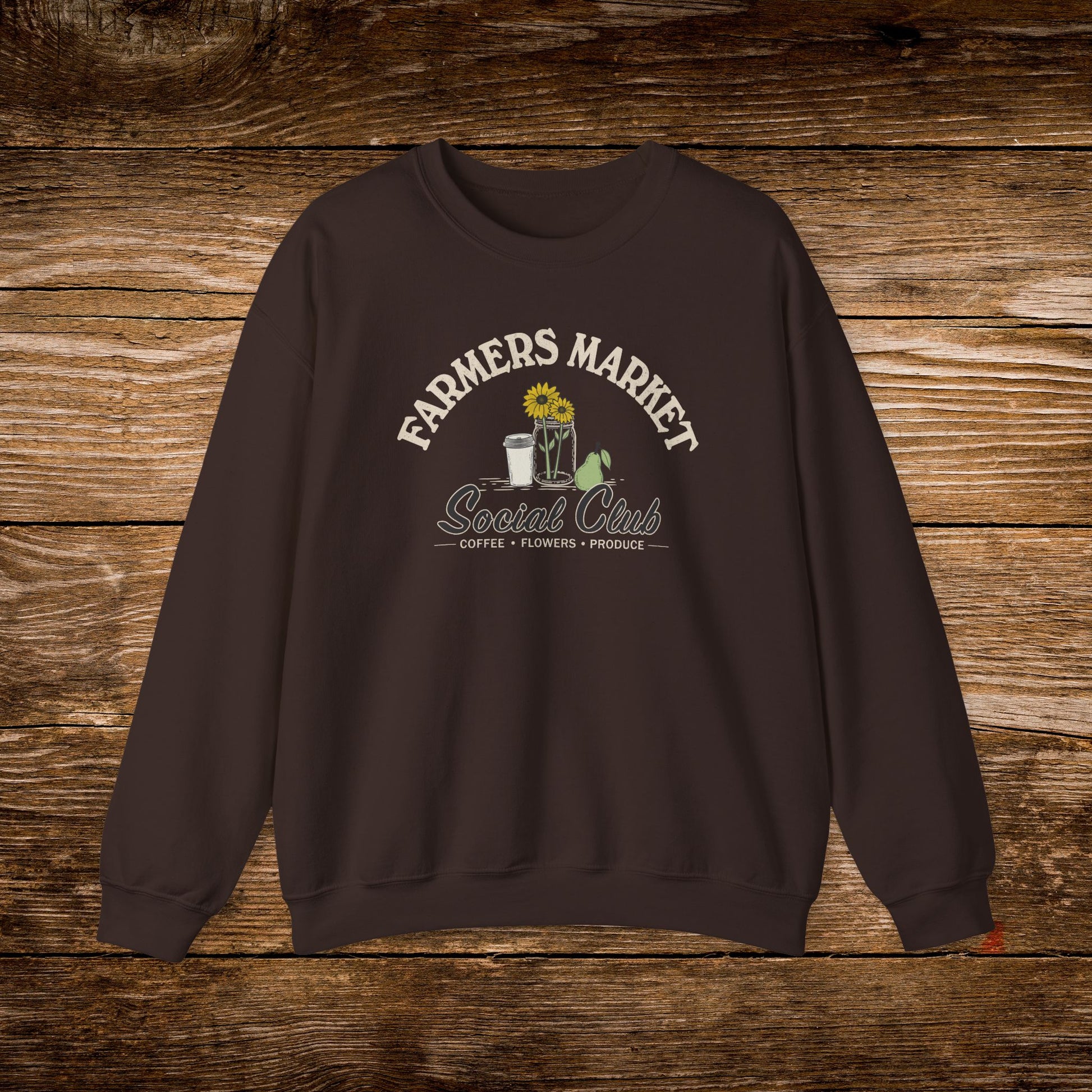 A dark brown crewneck sweatshirt with a graphic print reading 'FARMERS MARKET Social Club' and 'COFFEE • FRESHNESS • PROUD' beneath it, featuring a sunflower, coffee cup, and green leaves, displayed flat against a rustic wooden background.