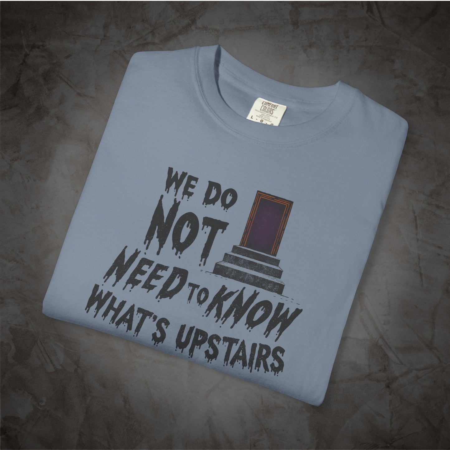 A folded light blue t-shirt with a dark gray graphic print that reads 'WE DO NOT NEED TO KNOW WHAT'S UPSTAIRS' alongside an illustration of a door, displayed on a textured gray background.