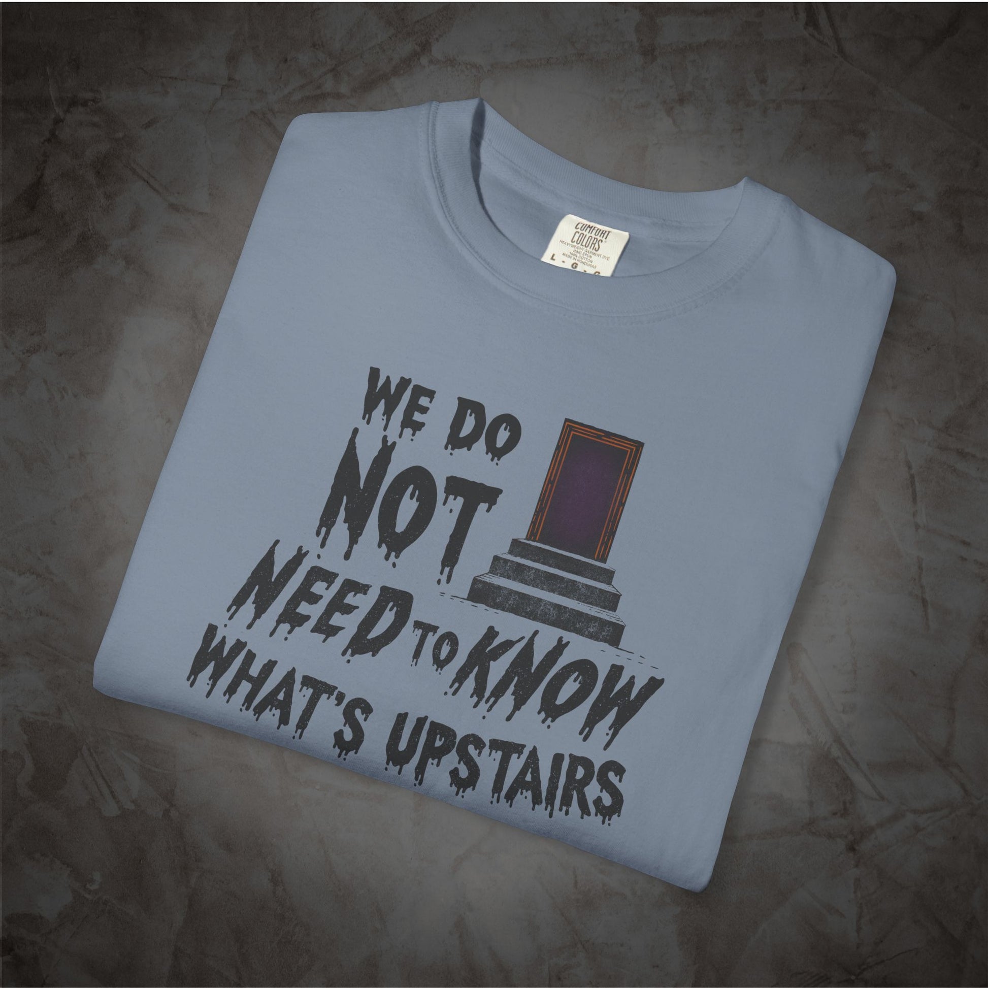A folded light blue t-shirt with a dark gray graphic print that reads 'WE DO NOT NEED TO KNOW WHAT'S UPSTAIRS' alongside an illustration of a door, displayed on a textured gray background.