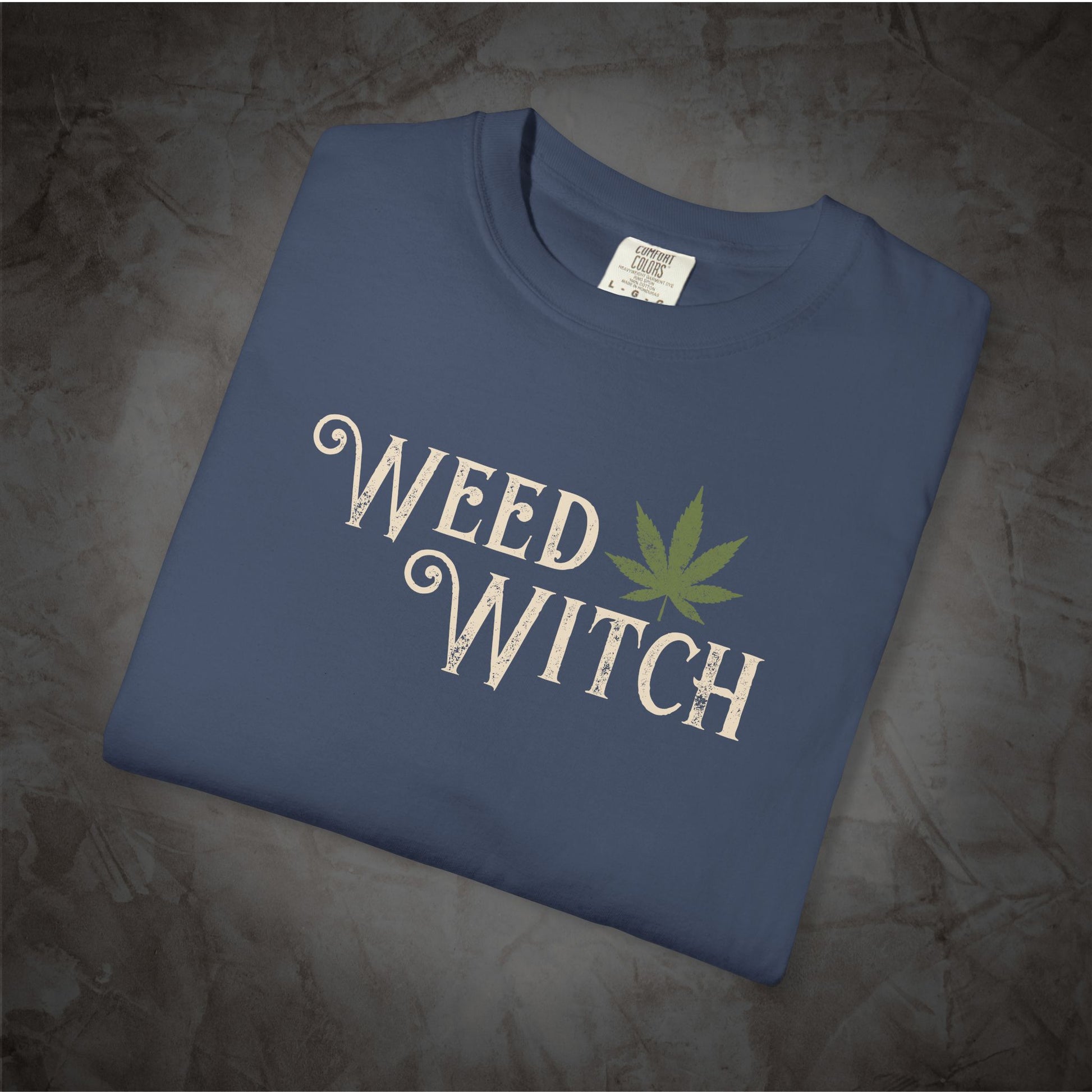 A folded slate blue t-shirt with a graphic print reading 'WEED WITCH' in a distressed white font, accented by a green cannabis leaf, displayed on a textured gray background.