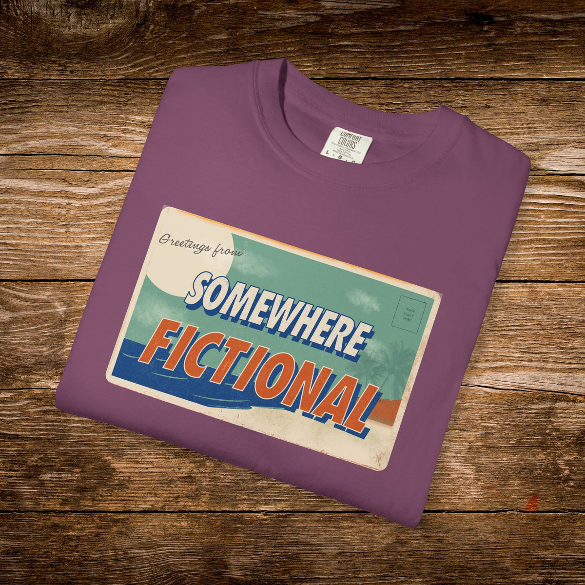 Folded Berry Comfort Colors t-shirt with the vintage “Greetings from Somewhere Fictional” postcard design.