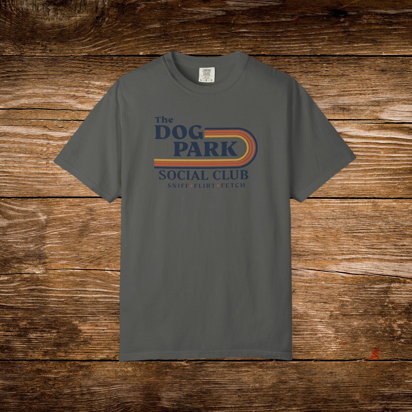 A dark gray crewneck t-shirt with a retro-style graphic reading 'The Dog Park Social Club' in purple and orange, laid flat on a rustic wooden plank background. The design text reads "Sniff, Flirt, Fetch."