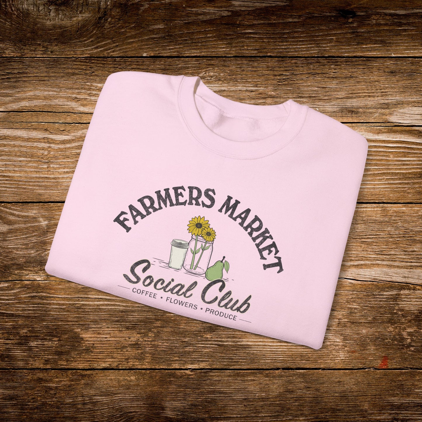 A light pink crewneck sweatshirt with a 'Farmers Market Social Club' graphic, folded neatly on a rustic wooden background.
