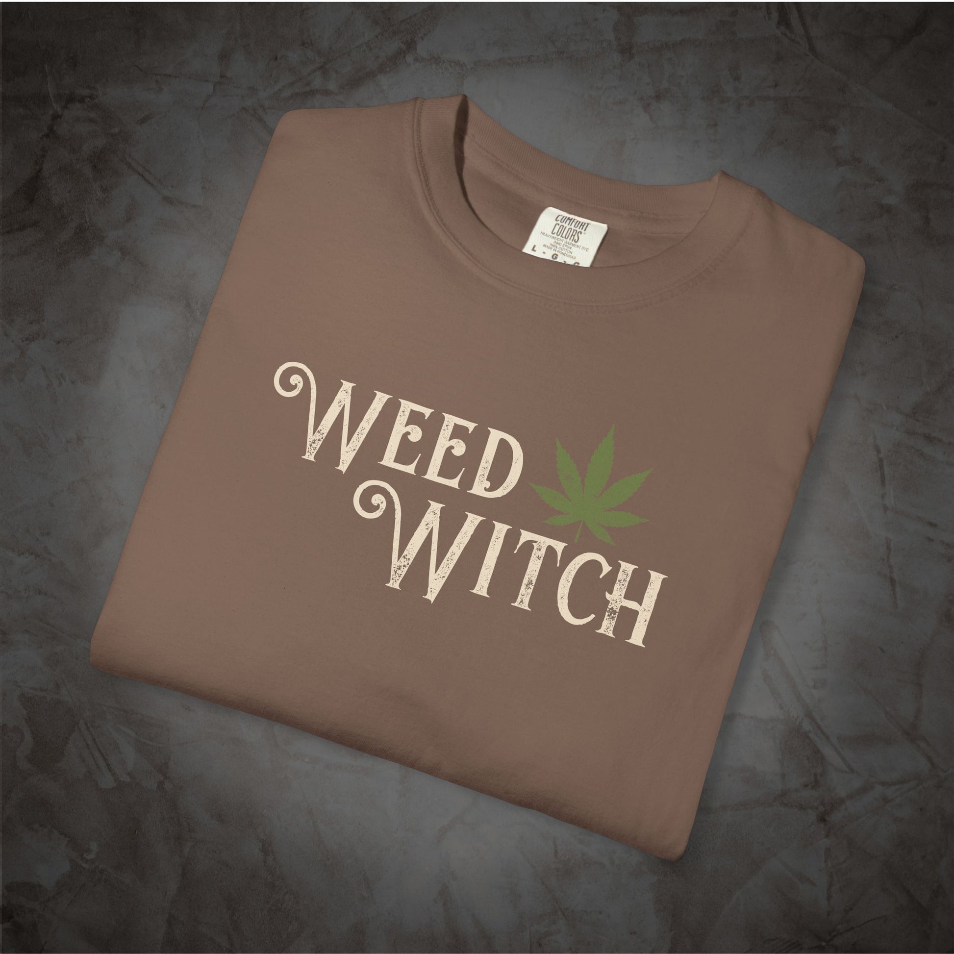 A brown folded t-shirt with a 'WEED WITCH' graphic in cream and a green cannabis leaf, displayed on a dark textured background.