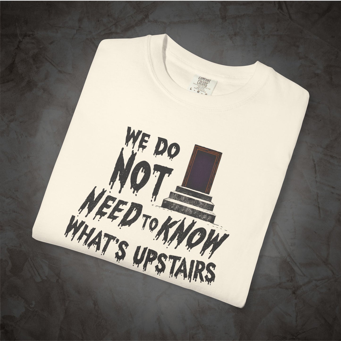 A folded off-white t-shirt with a black graphic print that reads 'WE DO NOT NEED TO KNOW WHAT'S UPSTAIRS' alongside a small illustration of a door, displayed on a dark textured background.