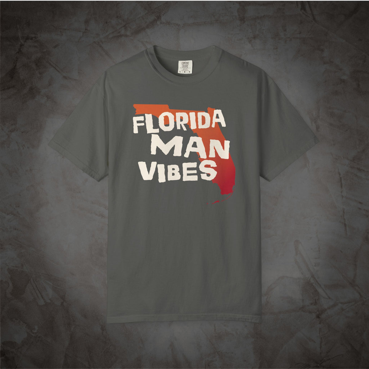 A dark gray t-shirt with a graphic print that reads 'FLORIDA MAN VIBES' in white letters over a red and orange Florida state silhouette, displayed against a textured gray background.