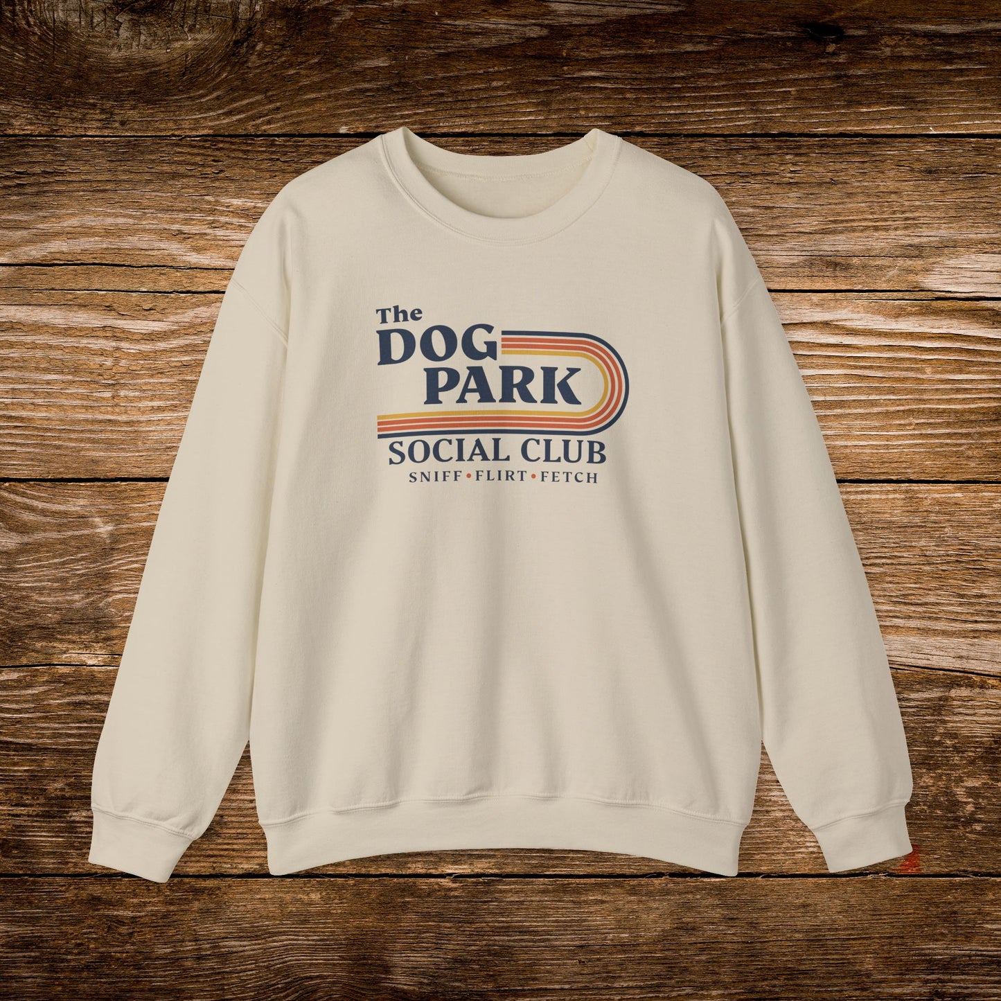 An off-white crewneck sweatshirt featuring a retro-style graphic that reads 'The DOG PARK SOCIAL CLUB' with 'SNIFF - FLIRT - FETCH' underneath, displayed flat against a rustic brown wooden plank background. The design text reads "Sniff, Flirt, Fetch."