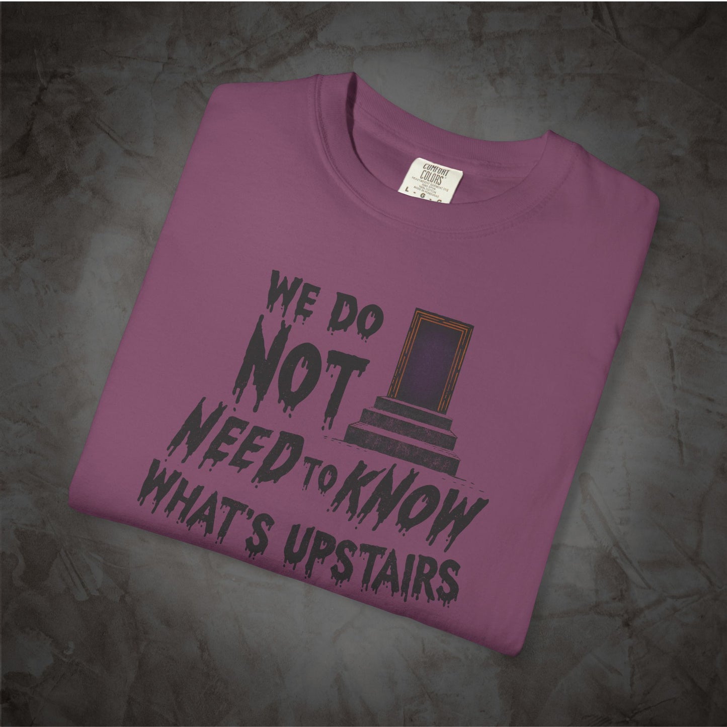 A folded mauve-purple t-shirt with a dark gray graphic print that reads 'WE DO NOT NEED TO KNOW WHAT'S UPSTAIRS' alongside an illustration of a door, displayed on a textured gray background.