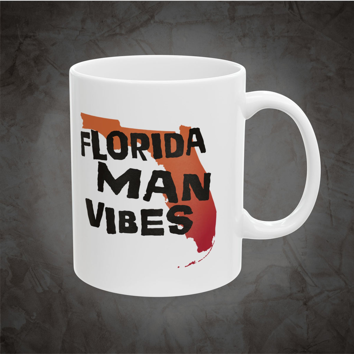 A white ceramic mug with a handle, featuring a graphic design of the state of Florida filled with orange and red brushstroke elements, with the words 'FLORIDA MAN VIBES' printed in black bold lettering, set against a dark textured gray background.