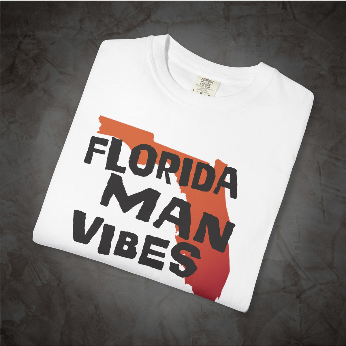 A folded white t-shirt with a graphic print of the state of Florida in orange and red, overlaid with the text 'FLORIDA MAN VIBES' in black. The t-shirt is displayed on a dark, textured gray background.