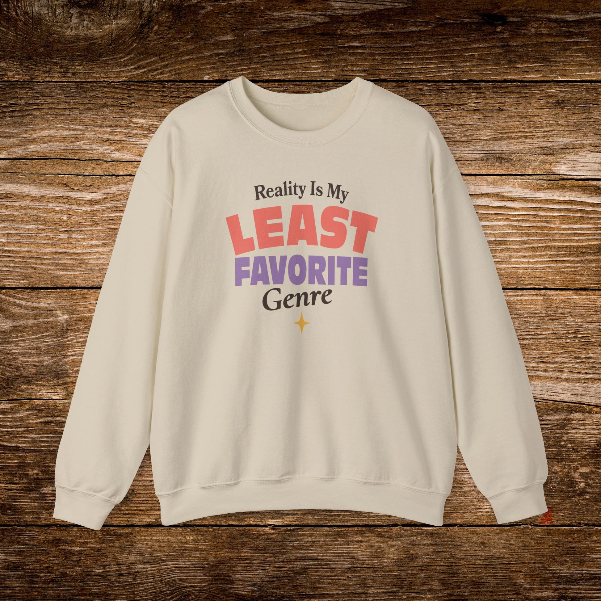 An off-white crewneck sweatshirt with a colorful graphic text print reading 'Reality Is My LEAST FAVORITE Genre' in red, purple, and black, displayed flat on a rustic brown wooden background.