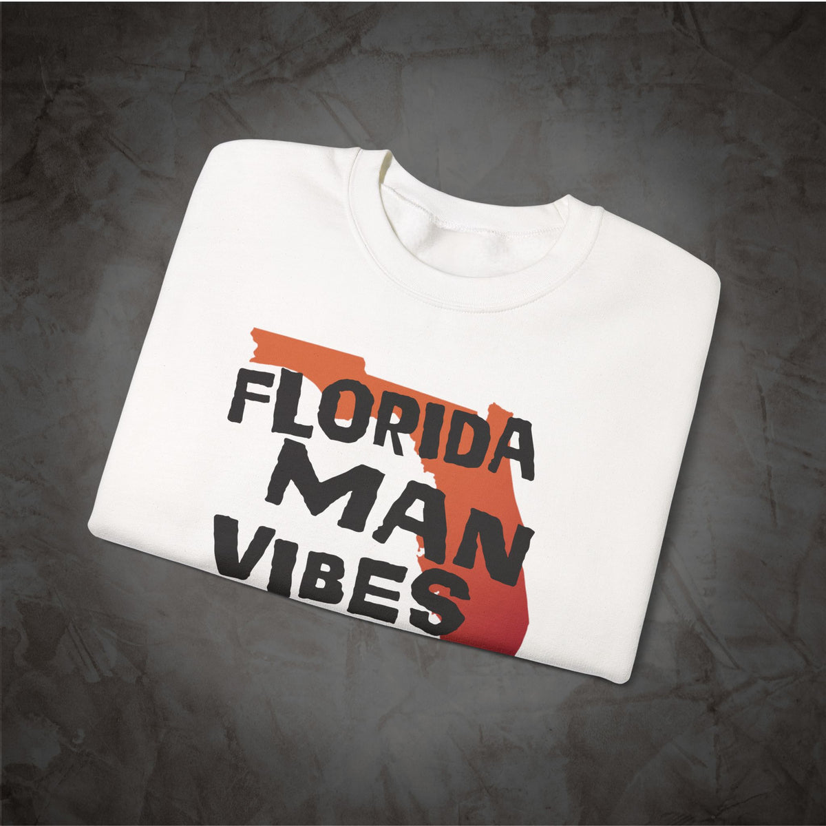 A folded white crewneck t-shirt with a graphic print featuring the words 'FLORIDA MAN VIBES' in black over an orange-to-red gradient silhouette of the state of Florida, displayed on a dark textured background.