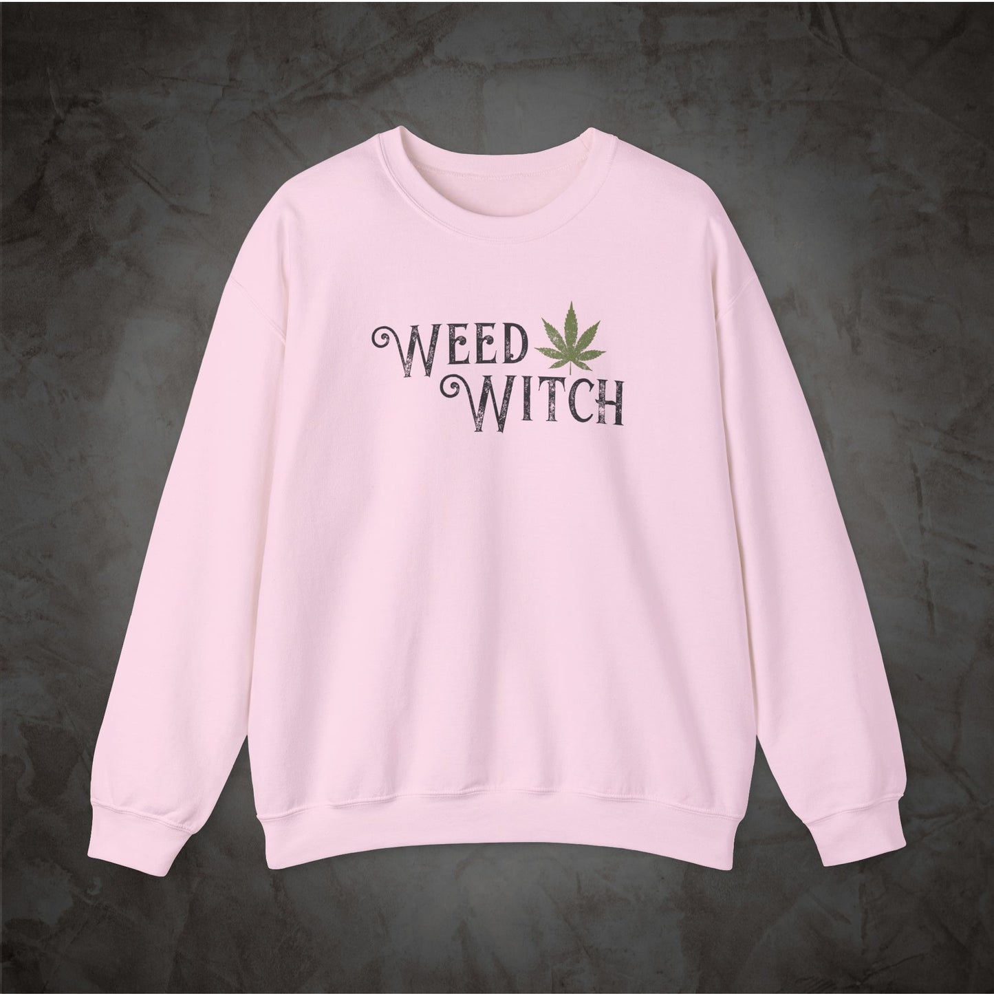 A light pink crewneck sweatshirt featuring the words 'WEED WITCH' in a dark gray gothic font with a green cannabis leaf graphic, displayed against a dark textured background.