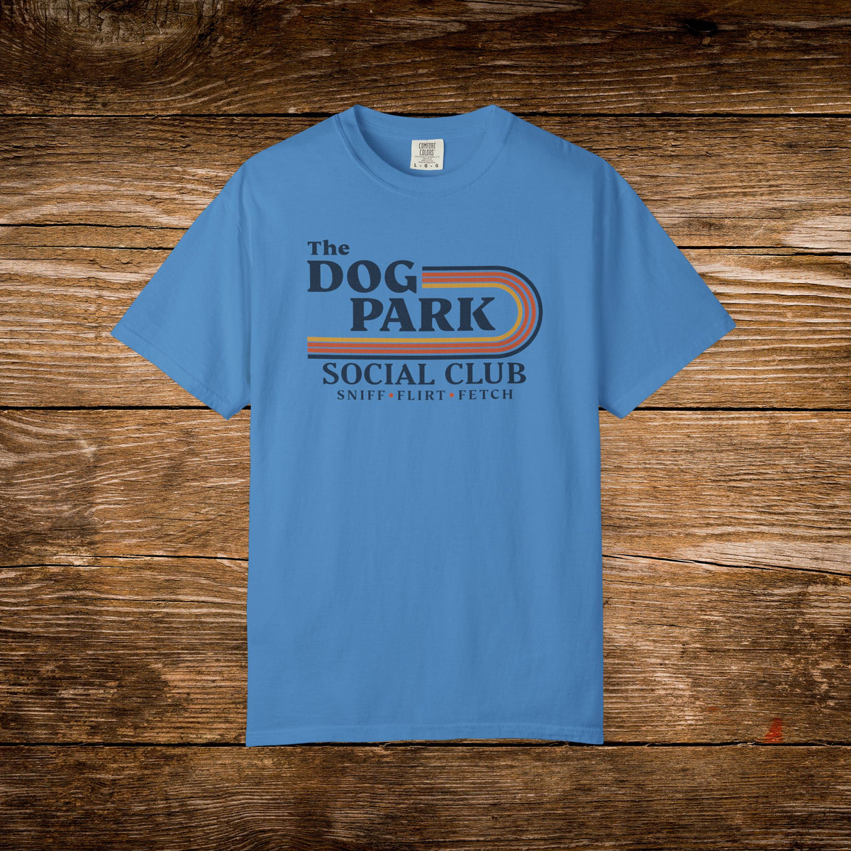 A light blue crewneck t-shirt with a retro-style graphic reading 'The DOG PARK SOCIAL CLUB' and 'SNIP-F-LIRT FETCH' in dark blue and orange, displayed flat against a rustic brown wooden plank background.