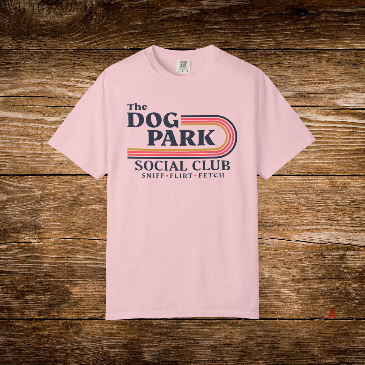 A light pink short-sleeve t-shirt with a retro-style graphic reading 'The Dog Park Social Club' and 'Sniff-Flirt-Fetch' in navy and orange-red tones, laid flat on a rustic wooden plank background.