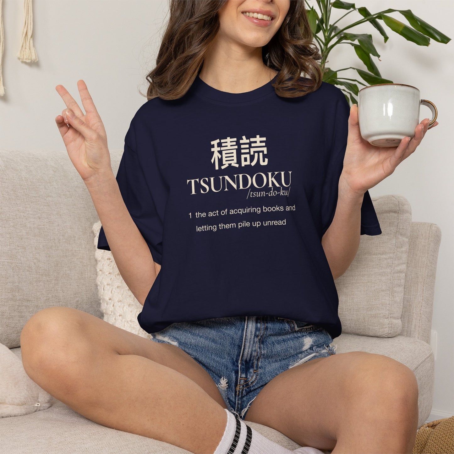 Person holding a mug while wearing a navy Tsundoku dictionary definition T-shirt