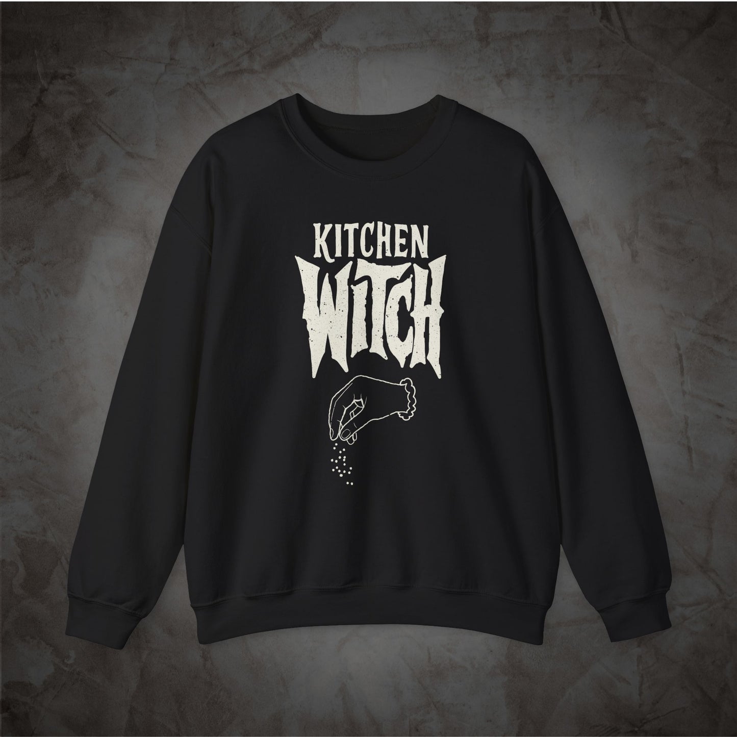 A black crewneck sweatshirt with a white graphic print reading 'KITCHEN WITCH' and a hand sprinkling seasoning, displayed against a textured gray background.