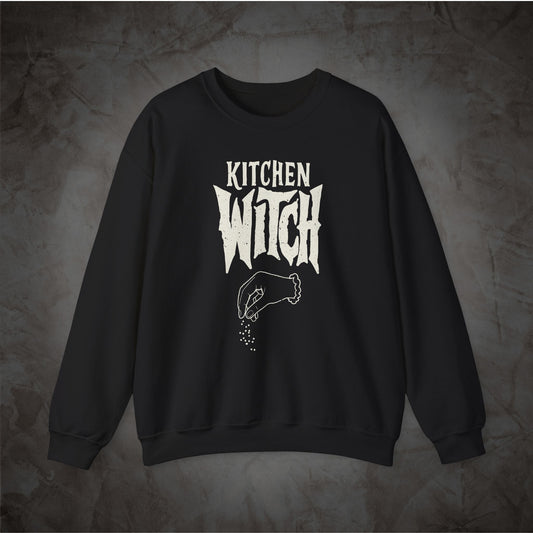 A black crewneck sweatshirt with a white graphic print reading 'KITCHEN WITCH' and a hand sprinkling seasoning, displayed against a textured gray background.