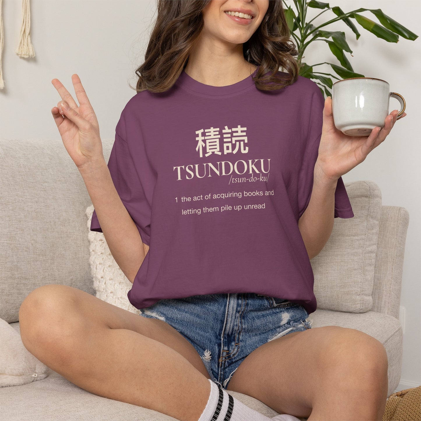 Person holding a mug while wearing a berry Tsundoku dictionary definition T-shirt.