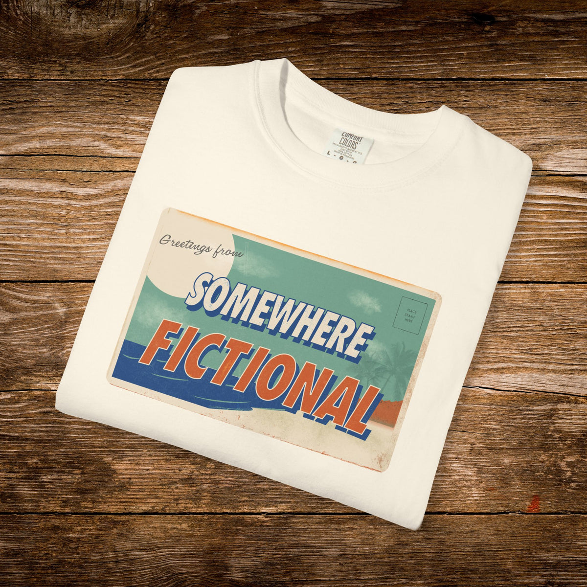Folded ivory Comfort Colors t-shirt displaying the “Greetings from Somewhere Fictional” vintage postcard design.