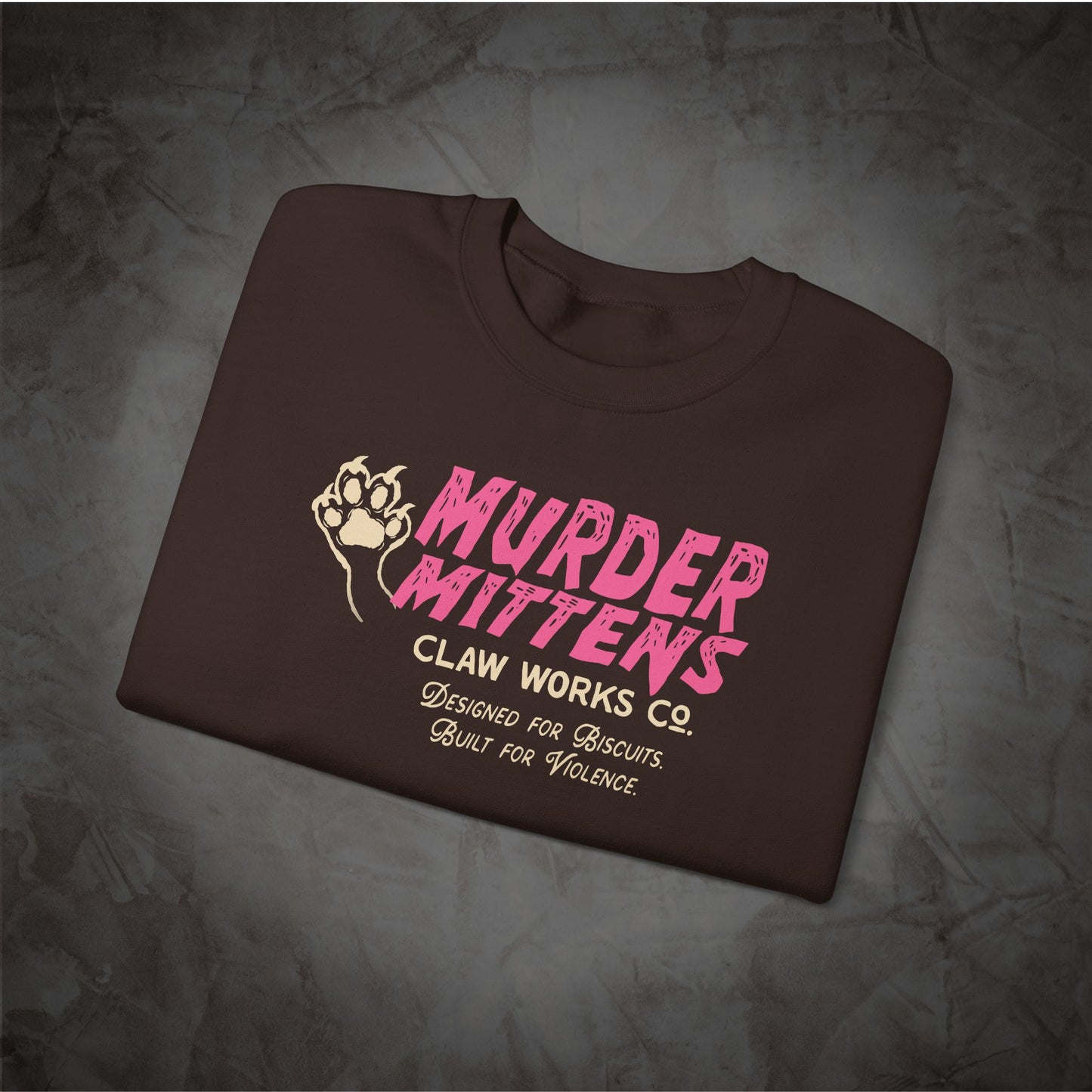 A dark brown folded t-shirt with a pink and beige graphic print reading 'MURDER MITTENS CLAW WORKS CO. DESIGNED FOR BISCUITS.