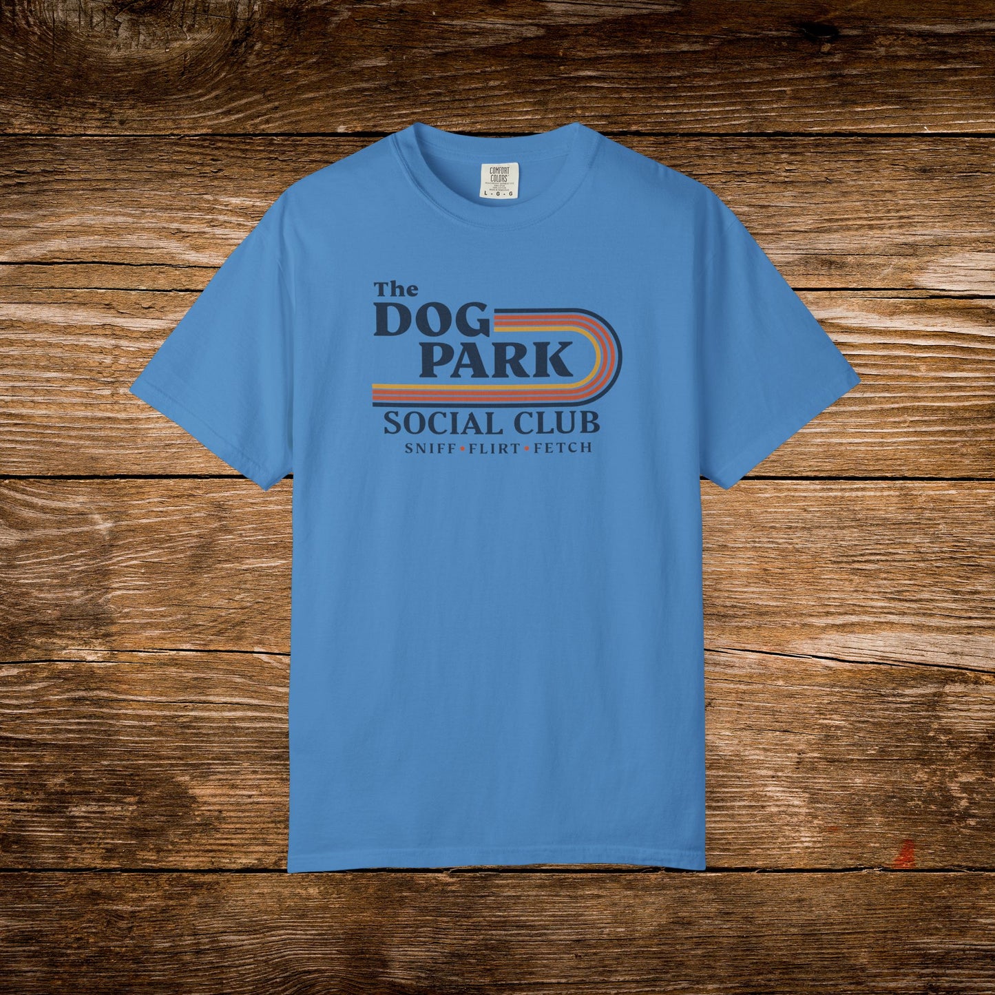 A light blue crewneck t-shirt featuring a retro-style graphic that reads 'The DOG PARK SOCIAL CLUB' with 'SNIFF FLIRT FETCH' underneath, displayed flat against a rustic brown wooden plank background. The design text reads "Sniff, Flirt, Fetch."