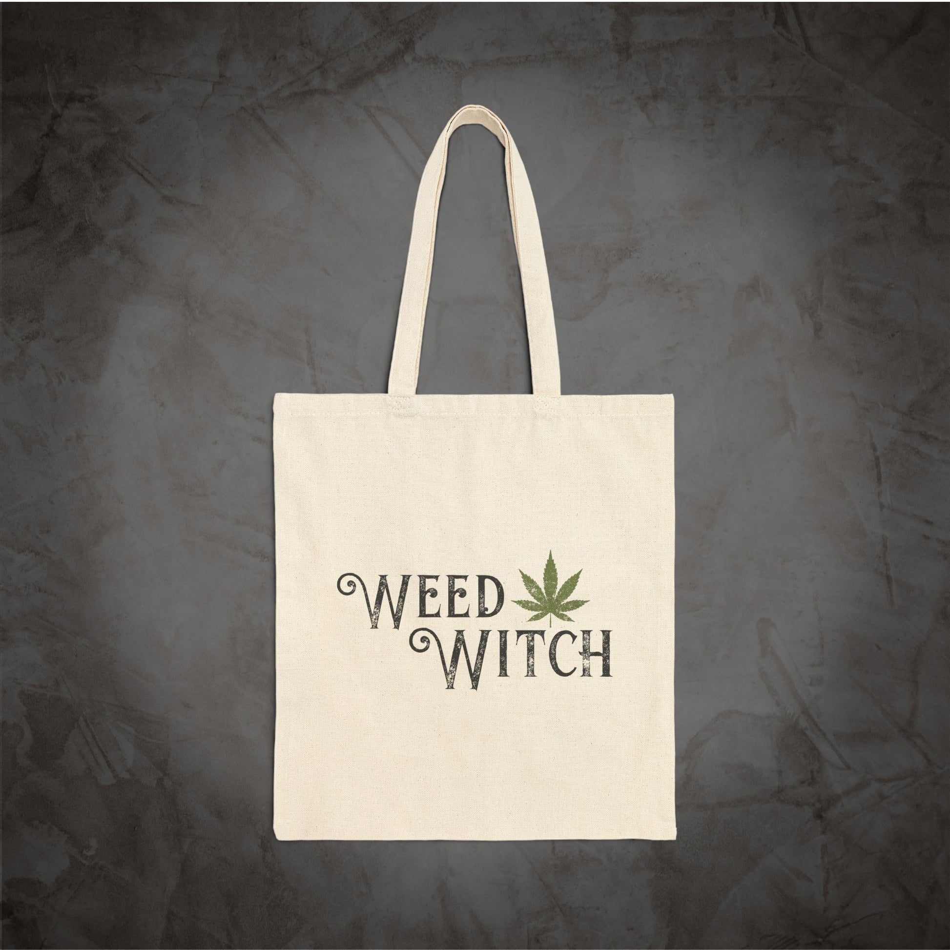 An off-white canvas tote bag with two handles, featuring the words 'WEED WITCH' in a dark gray gothic font and a green cannabis leaf graphic, set against a dark textured gray background.