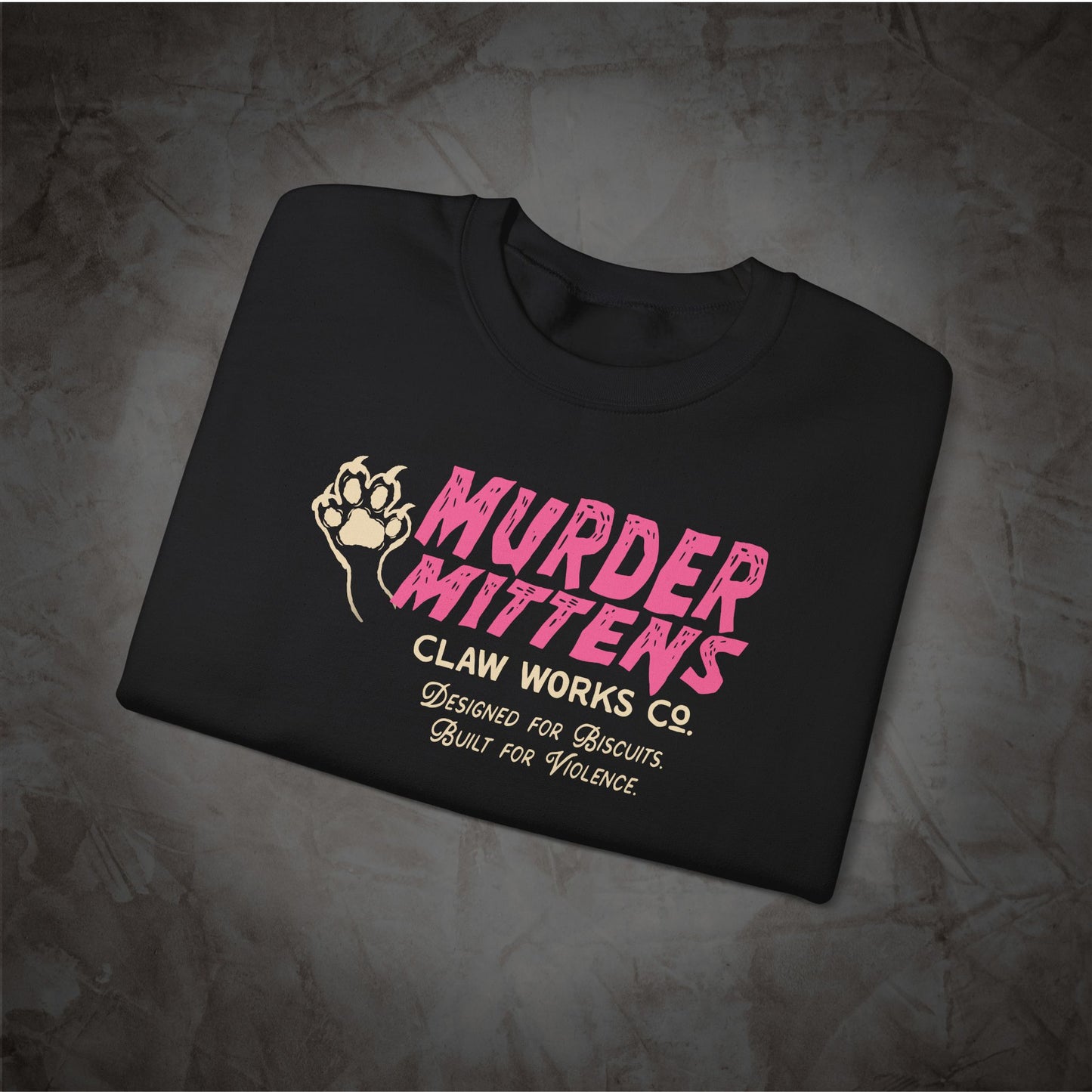 A black short-sleeve t-shirt laid flat on a textured gray background, featuring a pink and gold graphic print that reads 'MURDER MITTENS' with 'CLAW WORKS CO.' underneath.