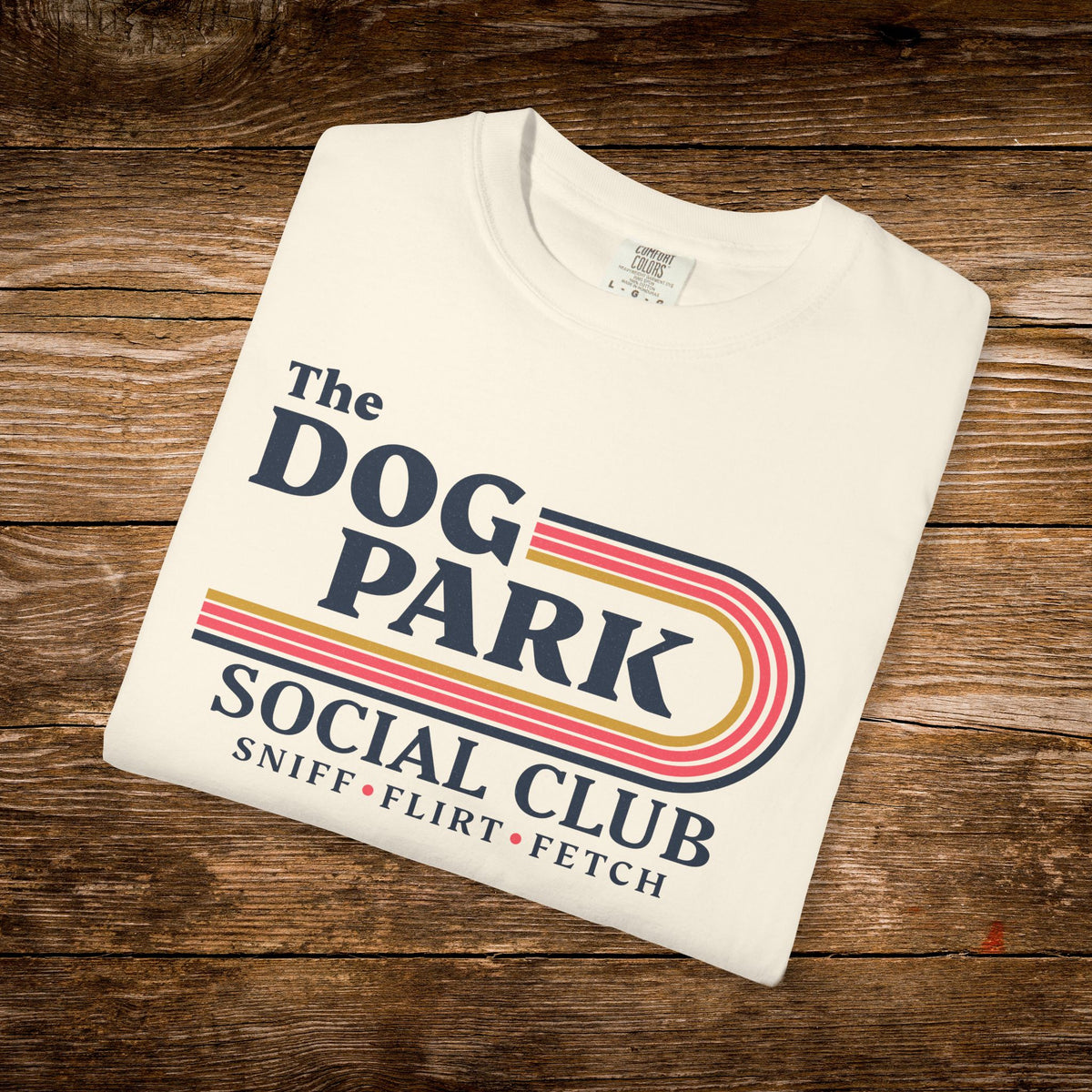 A folded off-white t-shirt with a retro-style 'The Dog Park Social Club' graphic, displayed on a rustic wooden plank background.